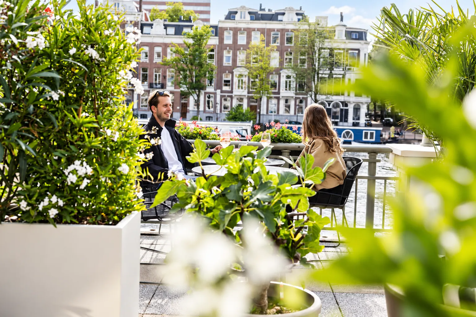 Gasten aan tafel tussen lichte plantenbakken met witte bloeiers, met uitzicht op de Amstel