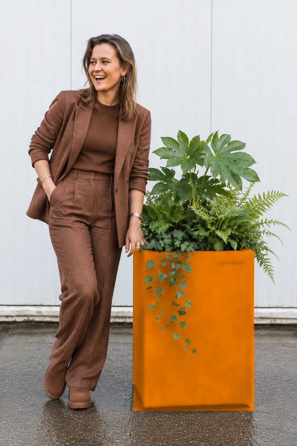 Lachende vrouw leunt tegen een oranje plantenbak met vingerplant, varens en klimop