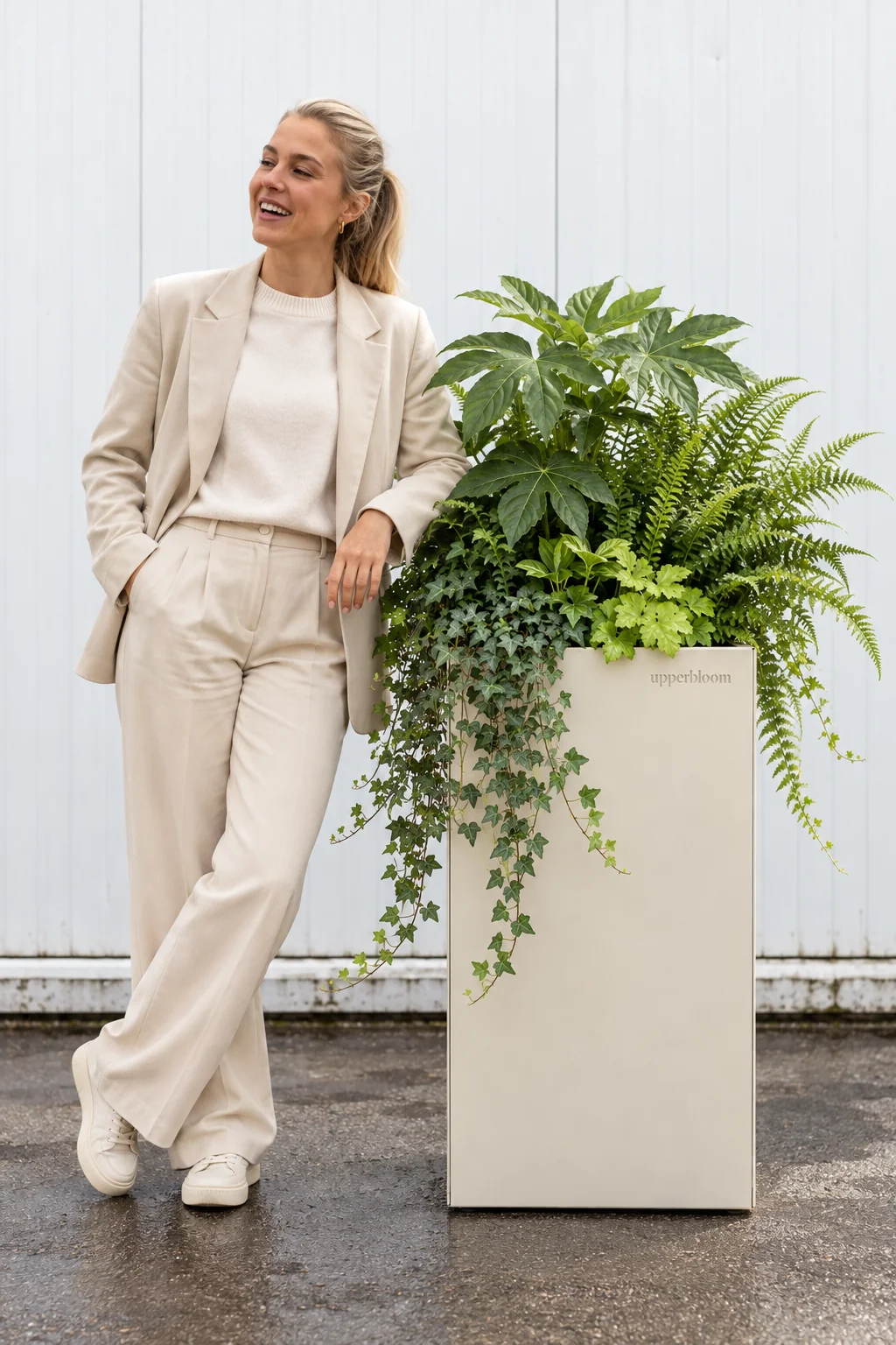 Lachende vrouw naast een hoge lichte plantenbak met vingerplant, varens en klimop