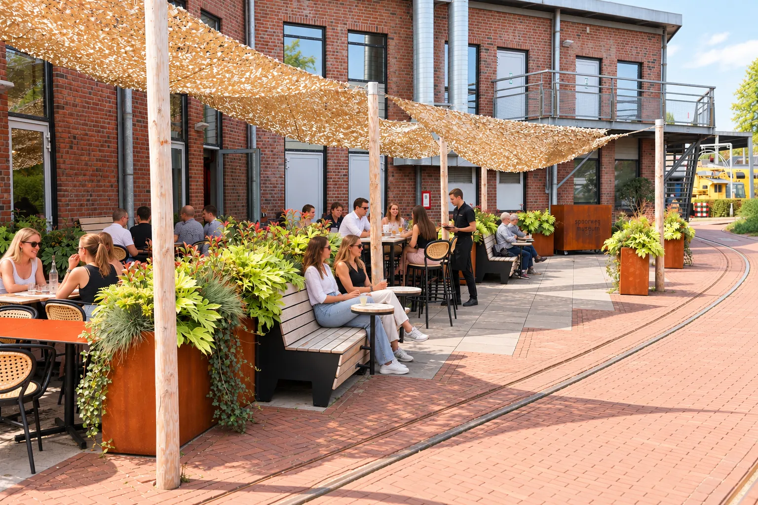 Terras bij een bakstenen gebouw met cortenstalen plantenbakken, banken en schaduwdoek boven gasten