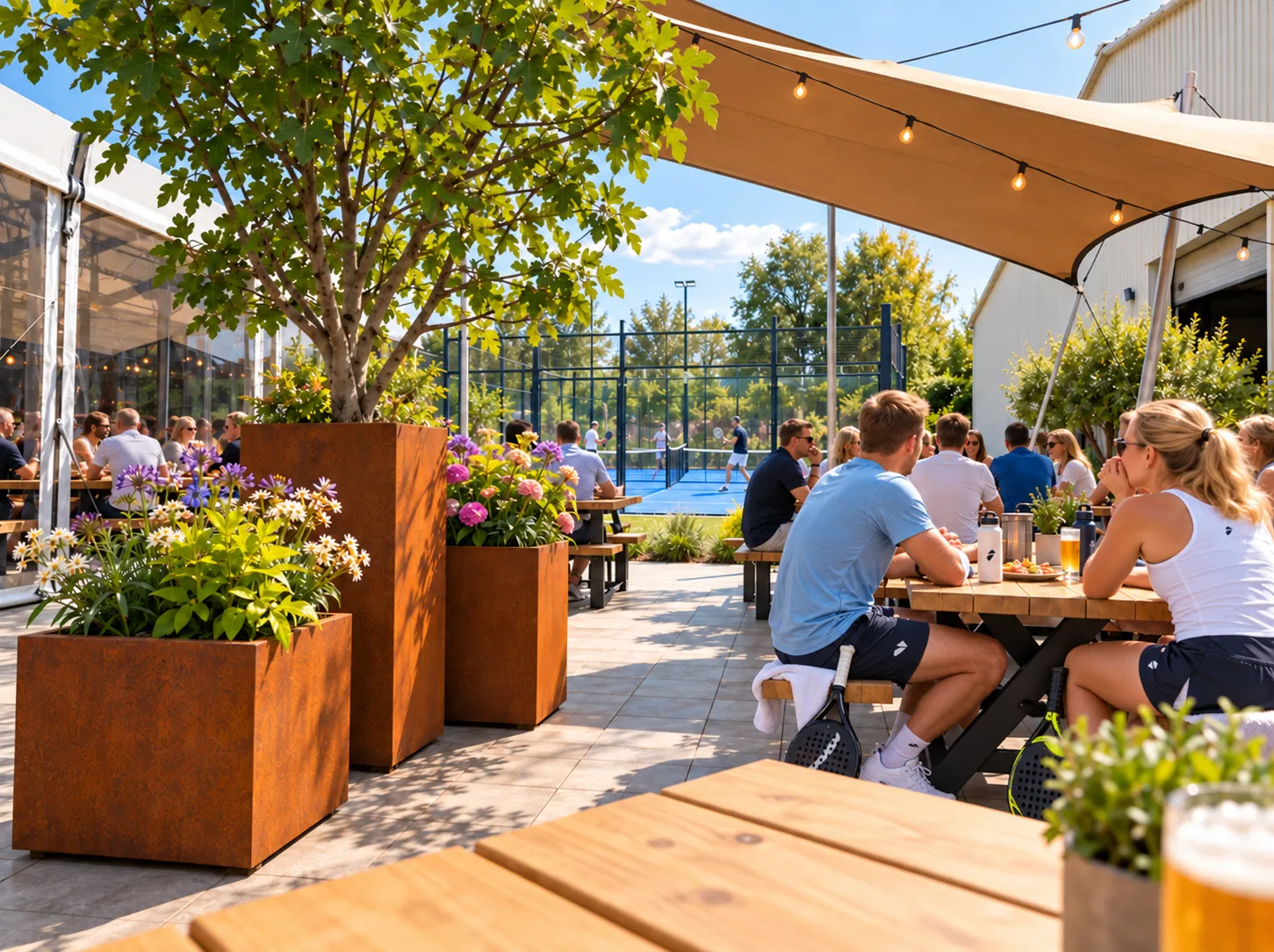 Cortenstalen plantenbakken met bloeiers en een boom naast de picknicktafels, met de blauwe padelbanen op de achtergrond
