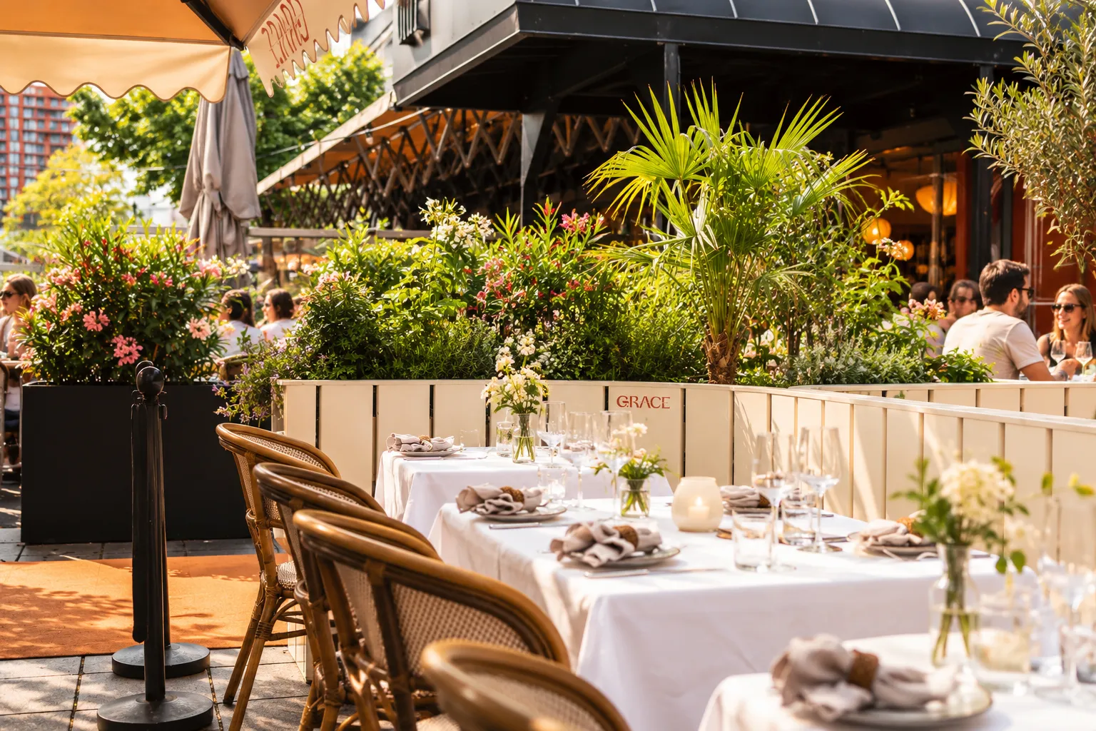 Gedekte tafels met witte tafelkleden en rieten stoelen langs de lichte plantenbakken van Grace, met een zwarte bak vol roze bloeiers en een palm erachter