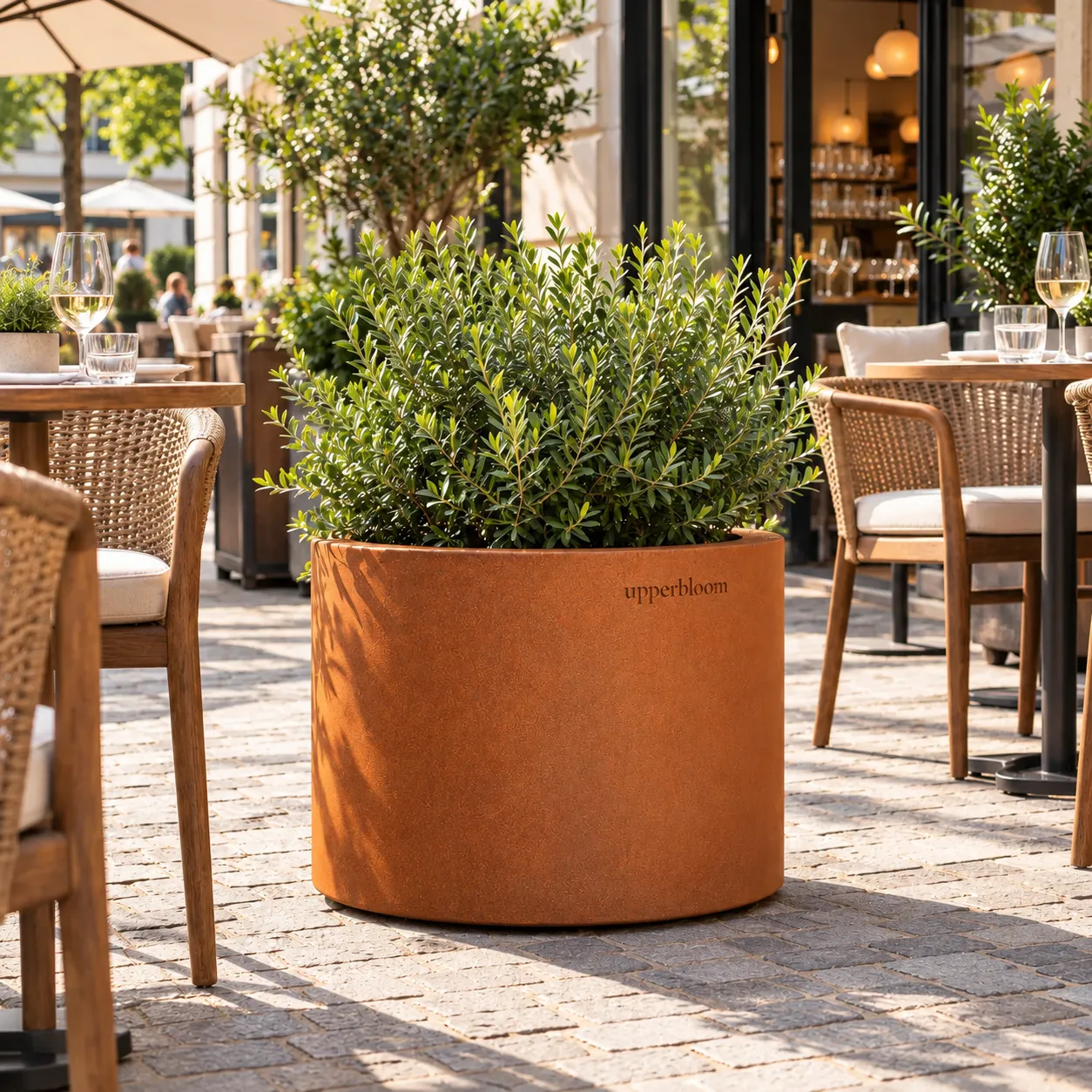 Ronde cortenstalen plantenbak met groenblijvende heester tussen rieten stoelen op een terras