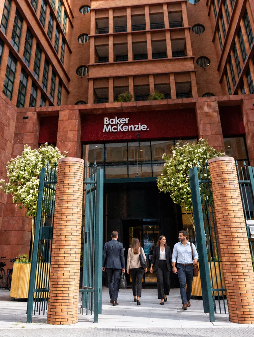 Entree van Baker McKenzie met open stalen poort, bakstenen pilaren en twee bloeiende bomen in houten plantenbakken