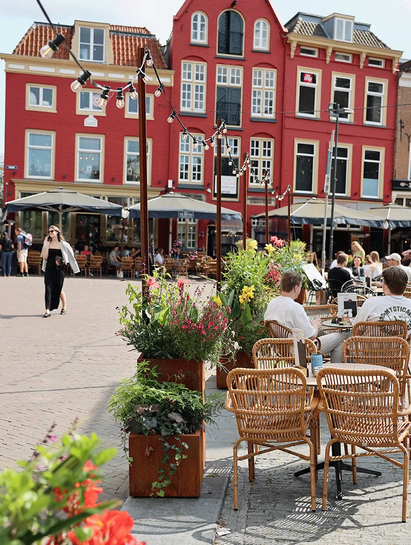 Twee cortenstalen bakken in verschillende hoogtes met bloeiers en klimop naast een terras met rieten stoelen, voor een rode gevel