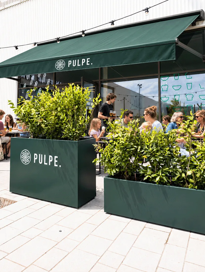 Donkergroene rechthoekige plantenbakken met het logo van Pulpe en heesters, voor het terras onder de groene luifel