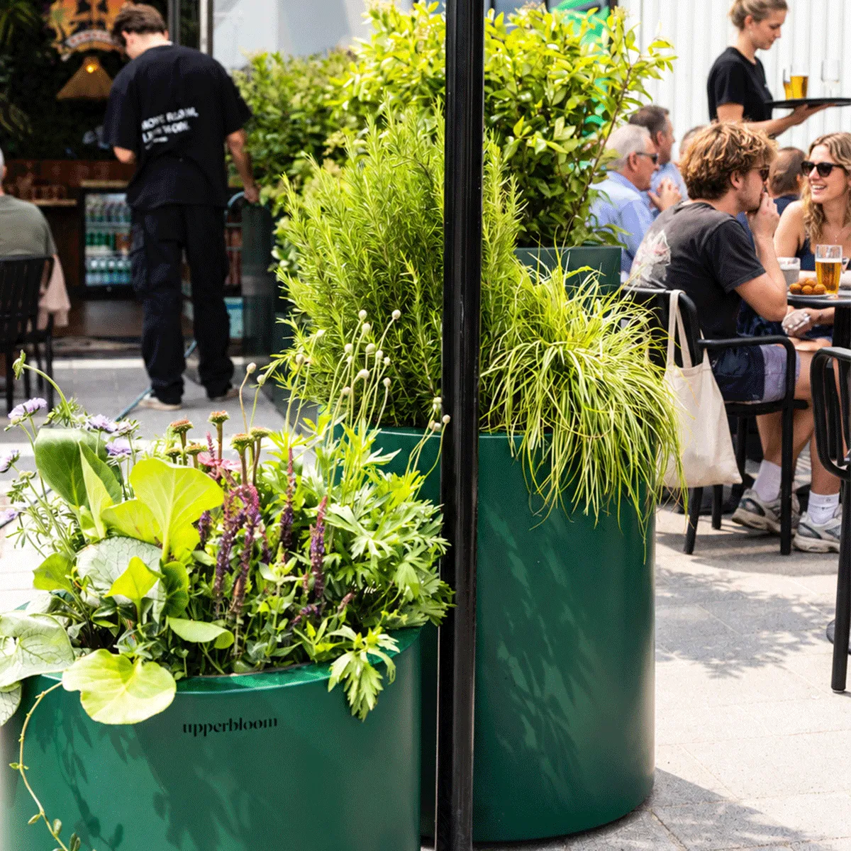 Donkergroene ronde plantenbakken met grassen, bloeiers en bladplanten tussen de tafels van het terras