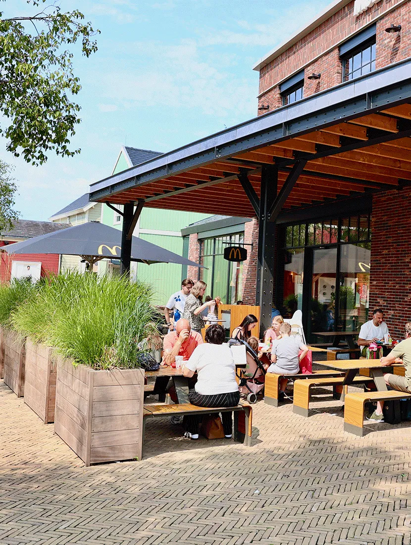Gasten op houten banken onder de overkapping van McDonald's, met hardhouten plantenbakken vol siergrassen langs het terras