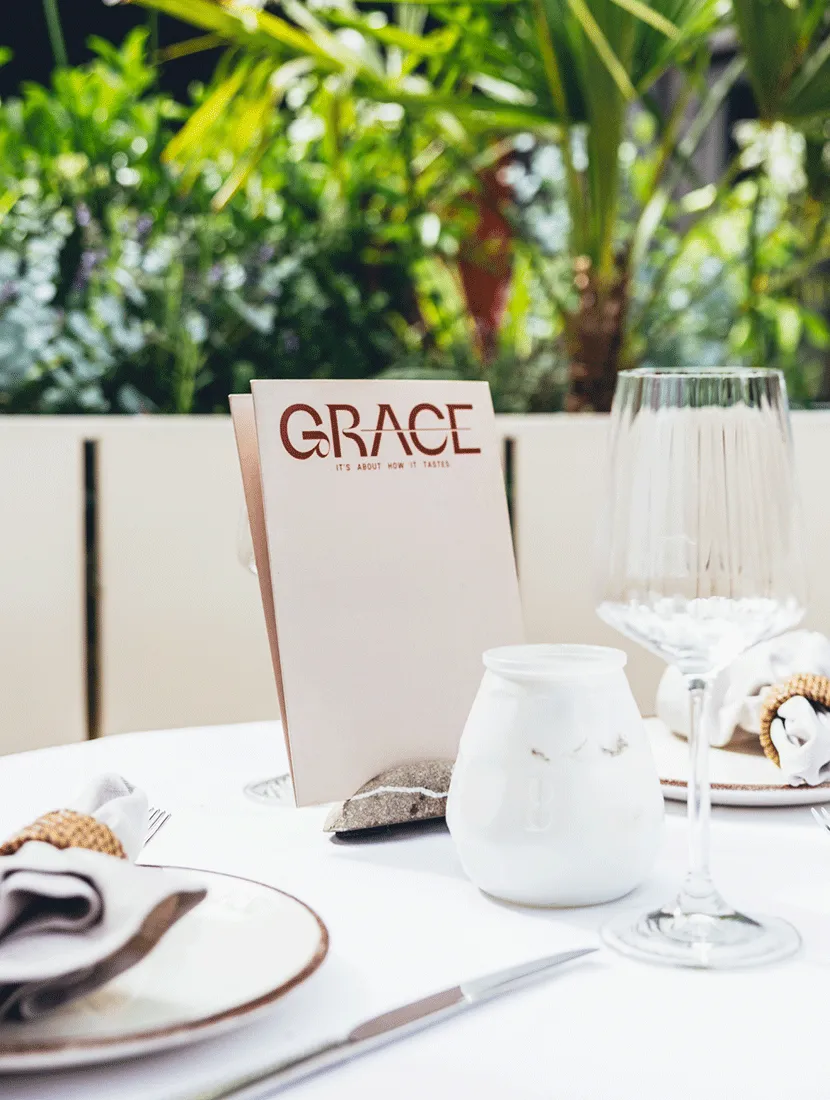 Menukaart van Grace op een gedekte tafel, met de lichte plantenbakken en het groen erachter