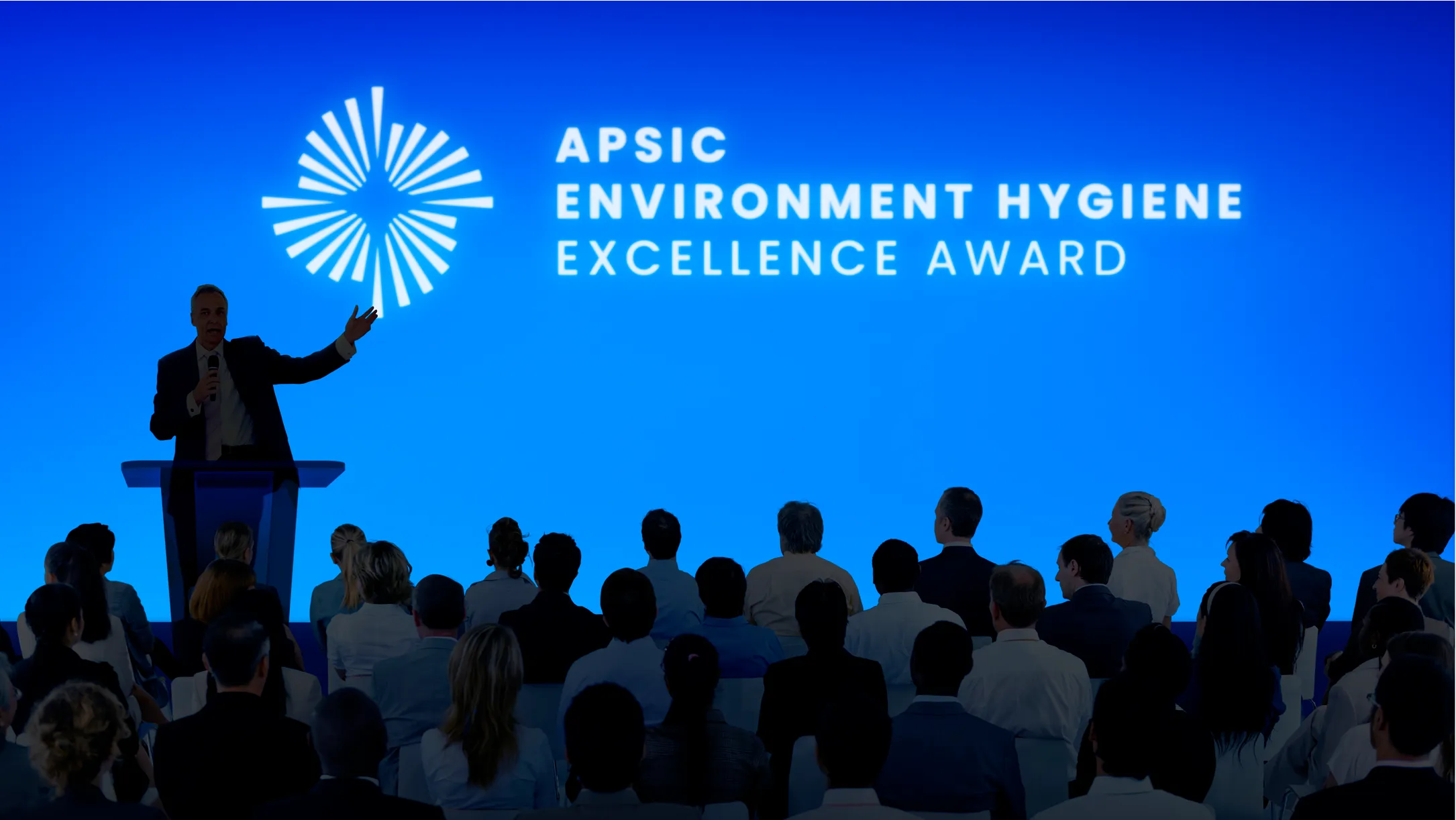 Speaker at podium addressing audience in front of blue screen with APSIC Environment Hygiene Excellence Award text.