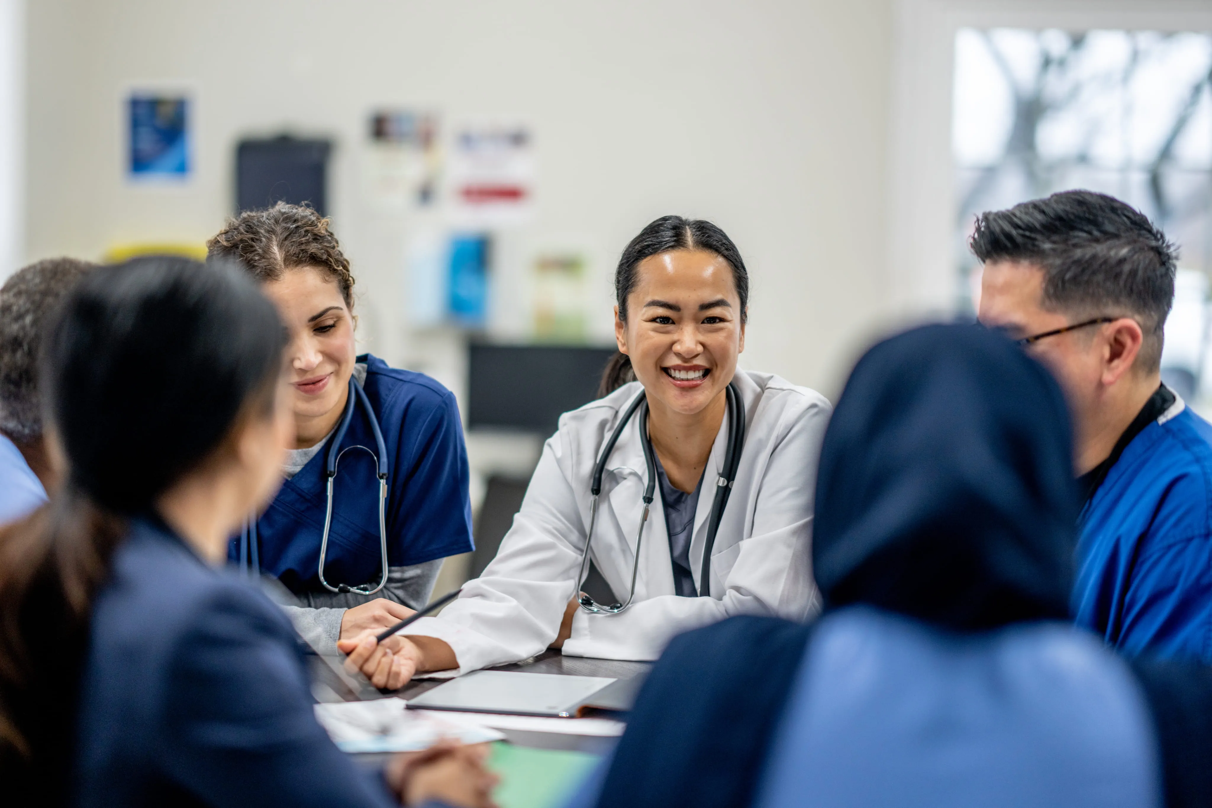 Smiling medical professionals wearing scrubs and lab coats in a group meeting in a bright room.