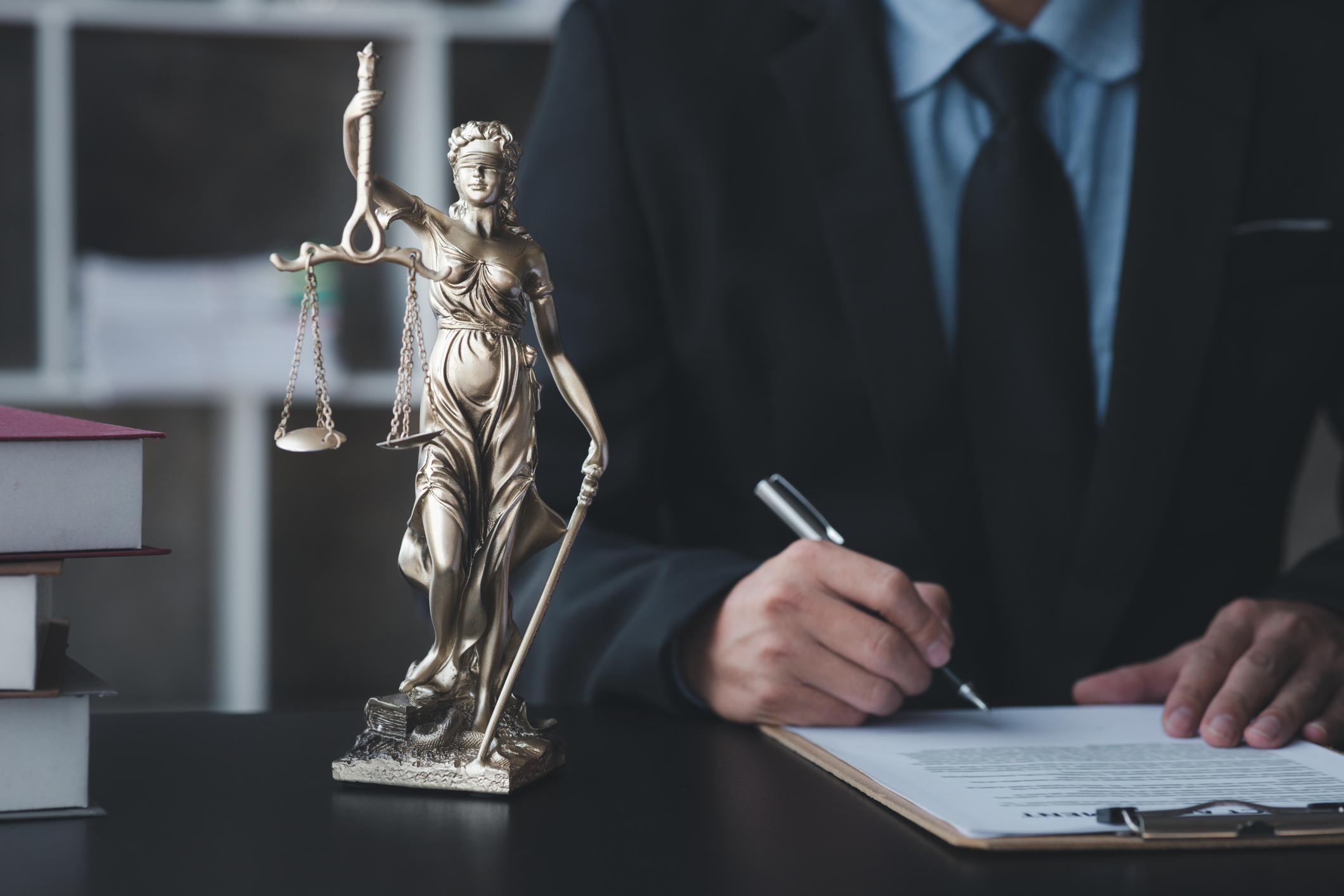 Blindfolded Lady Justice statue holding scales and sword on desk with man signing documents.