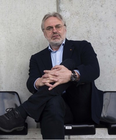 Man with glasses in a black suit sitting cross-legged on stadium seats against a concrete wall.