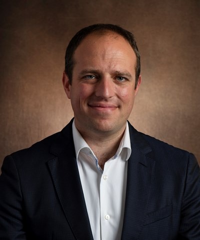 Smiling man in a dark suit jacket and white shirt against a brown background.