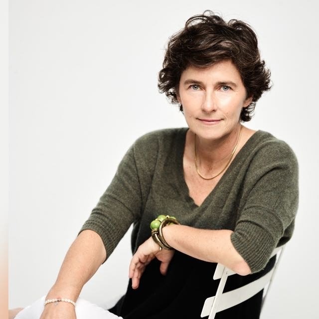 Woman with short curly hair wearing a green sweater and bracelets, seated on a white chair, looking at camera.