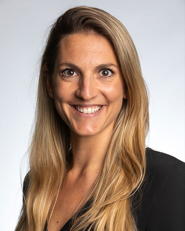 Smiling woman with long blonde hair wearing a black top against a plain light background.
