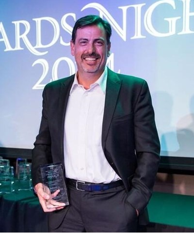 Man in black suit holding an award, smiling, standing in front of a screen with text 'AWARDS NIGHT 2024'.