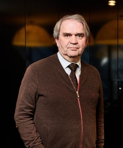 Middle-aged man with gray hair wearing a brown sweater and tie, standing against a dark background.