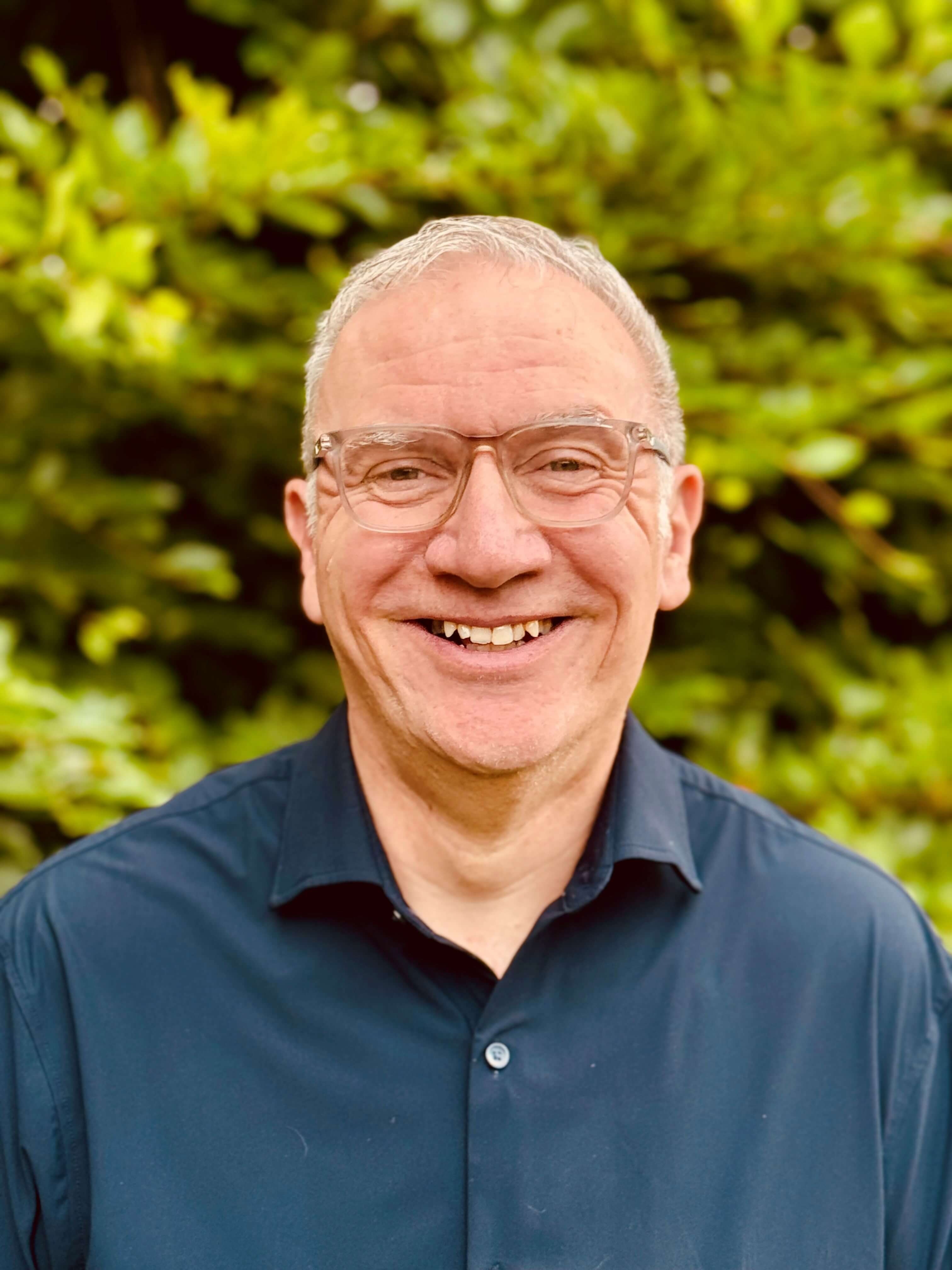 Smiling man with glasses wearing a dark blue shirt, standing in front of green foliage.