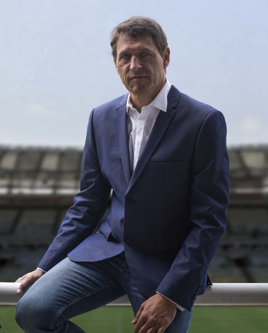 Man in a blue blazer and jeans sitting on a railing with a stadium blurred in the background.