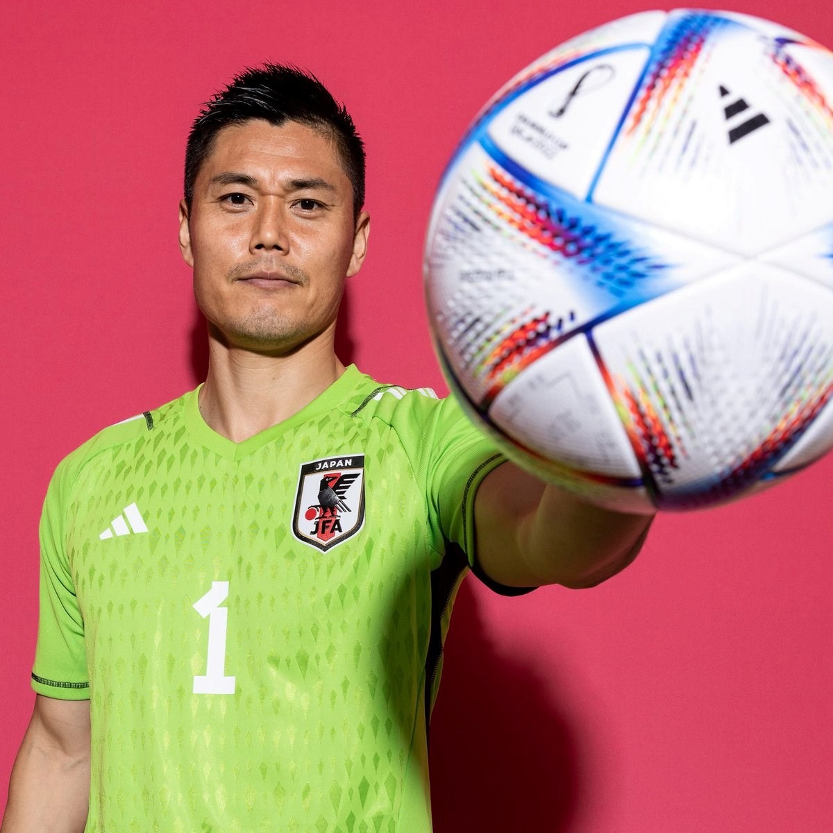 Man in green Japan soccer jersey holding a colorful soccer ball toward the camera on a red background.