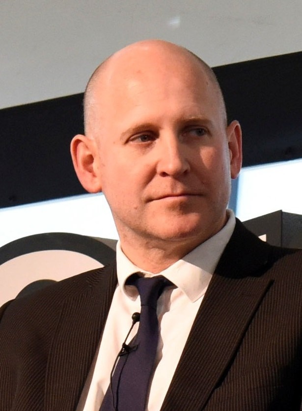Bald man in a dark suit, white shirt, and dark tie looking to the side while seated indoors.