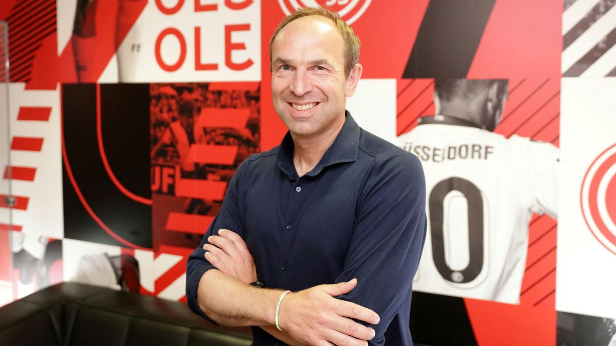 Smiling man in a navy shirt stands with arms crossed in front of a red and black sports-themed wall.