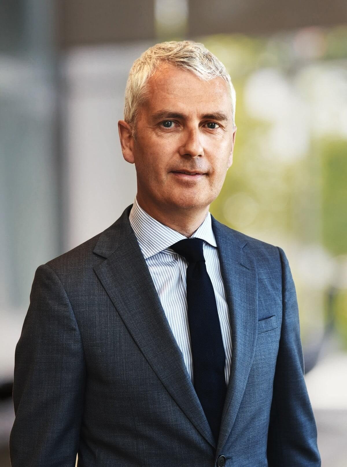 Middle-aged man with short gray hair wearing a dark suit, striped shirt, and black tie, looking at the camera.