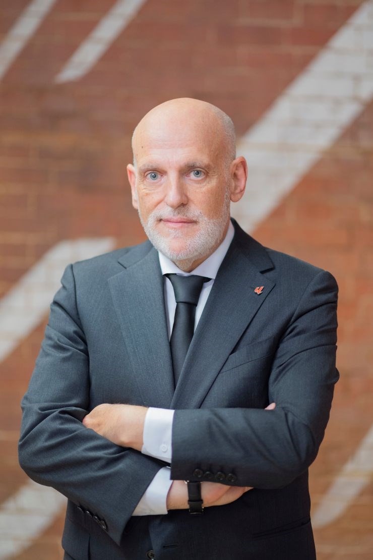 Bald man with gray beard in a dark suit and tie standing with arms crossed against brick wall.