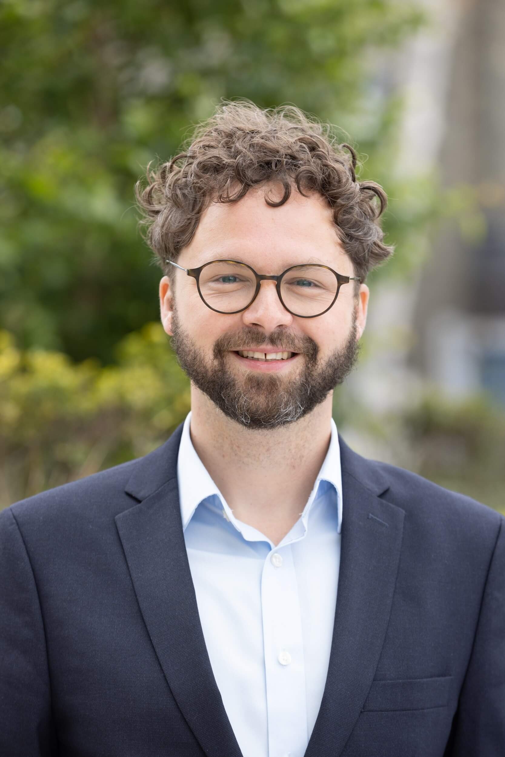 Smiling man with curly hair, beard, glasses wearing navy blazer and light blue shirt outdoors.
