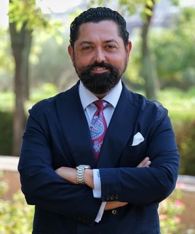 Man with beard in navy suit and paisley tie standing outdoors with arms crossed and smiling.