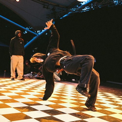 A breakdancer in a black hoodie and dark pants performing a dynamic move on a checkered floor with another person watching in the background under stage lighting.