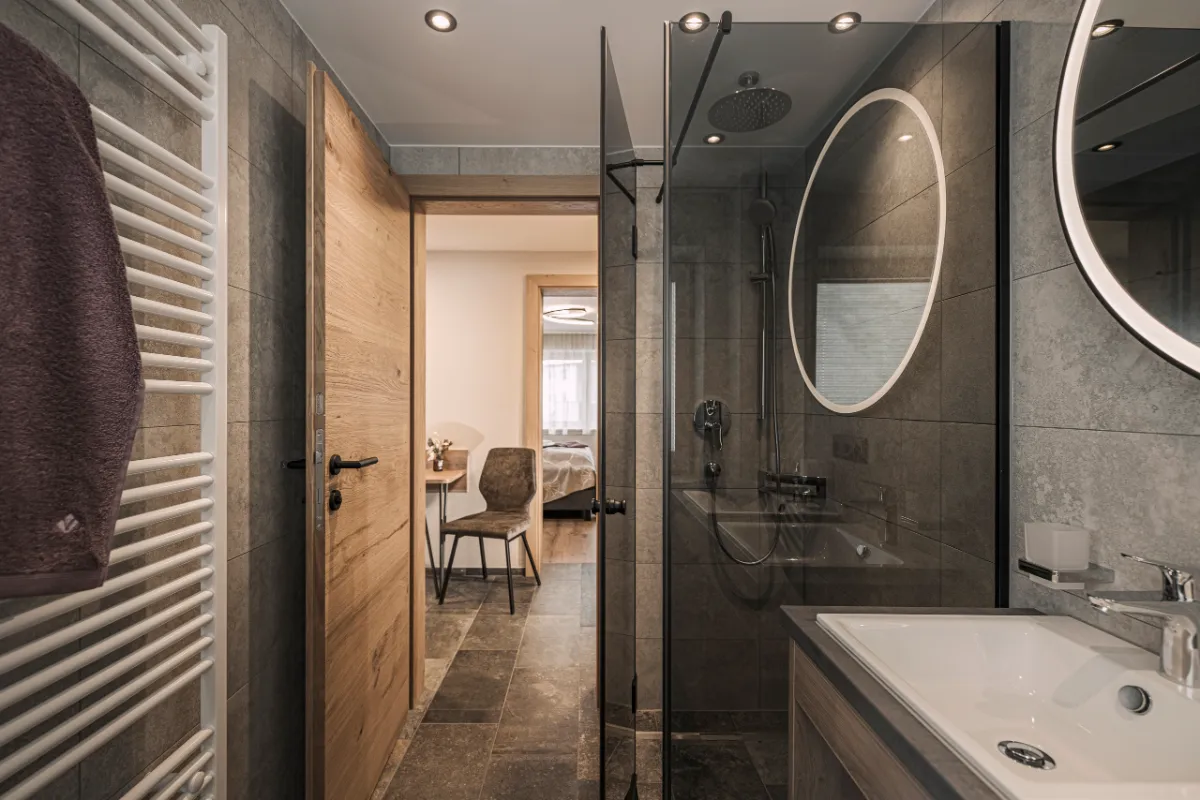Badezimmer im Ferienhaus Evelyn mit begehbarer Dusche, rundem Spiegel und Blick ins Schlafzimmer