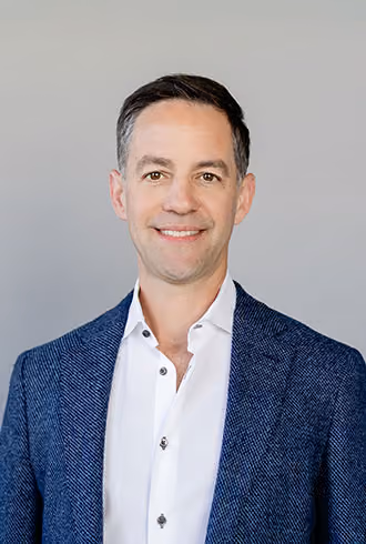 A man with short dark hair and a friendly expression wearing a white shirt and a blue blazer against a plain light gray background.