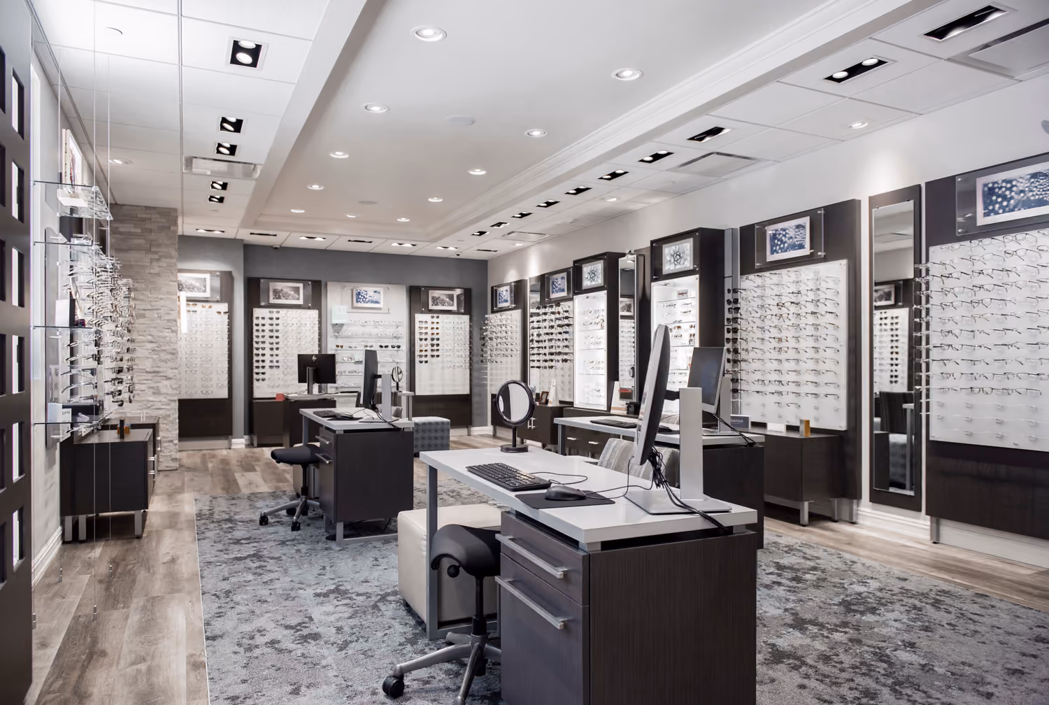 Modern optical store showroom with multiple display panels of eyeglass frames on walls, desks with computers and chairs on a patterned carpet, and bright overhead lighting.