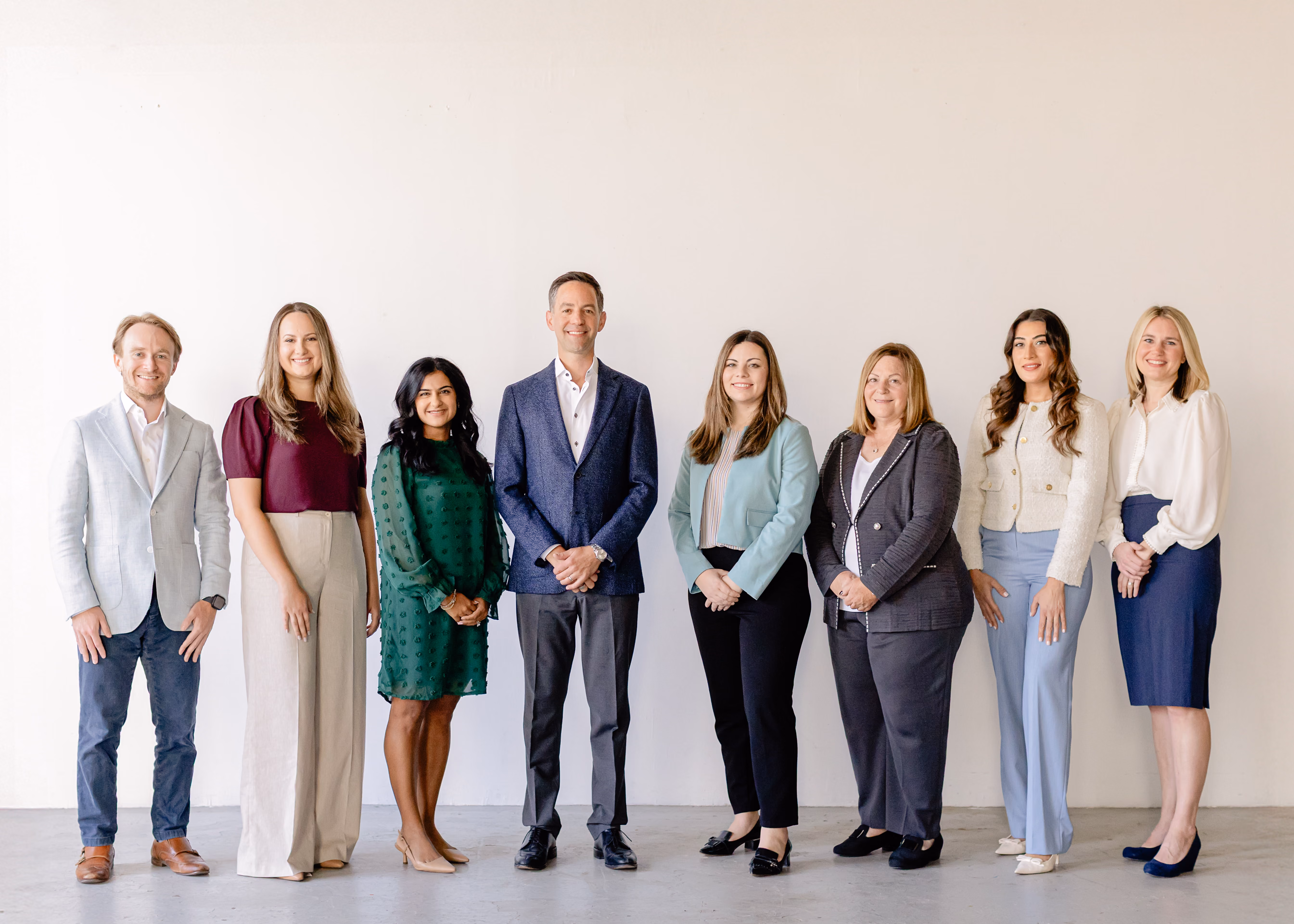 Group of eight professionally dressed people standing side by side against a plain light background, smiling at the camera.