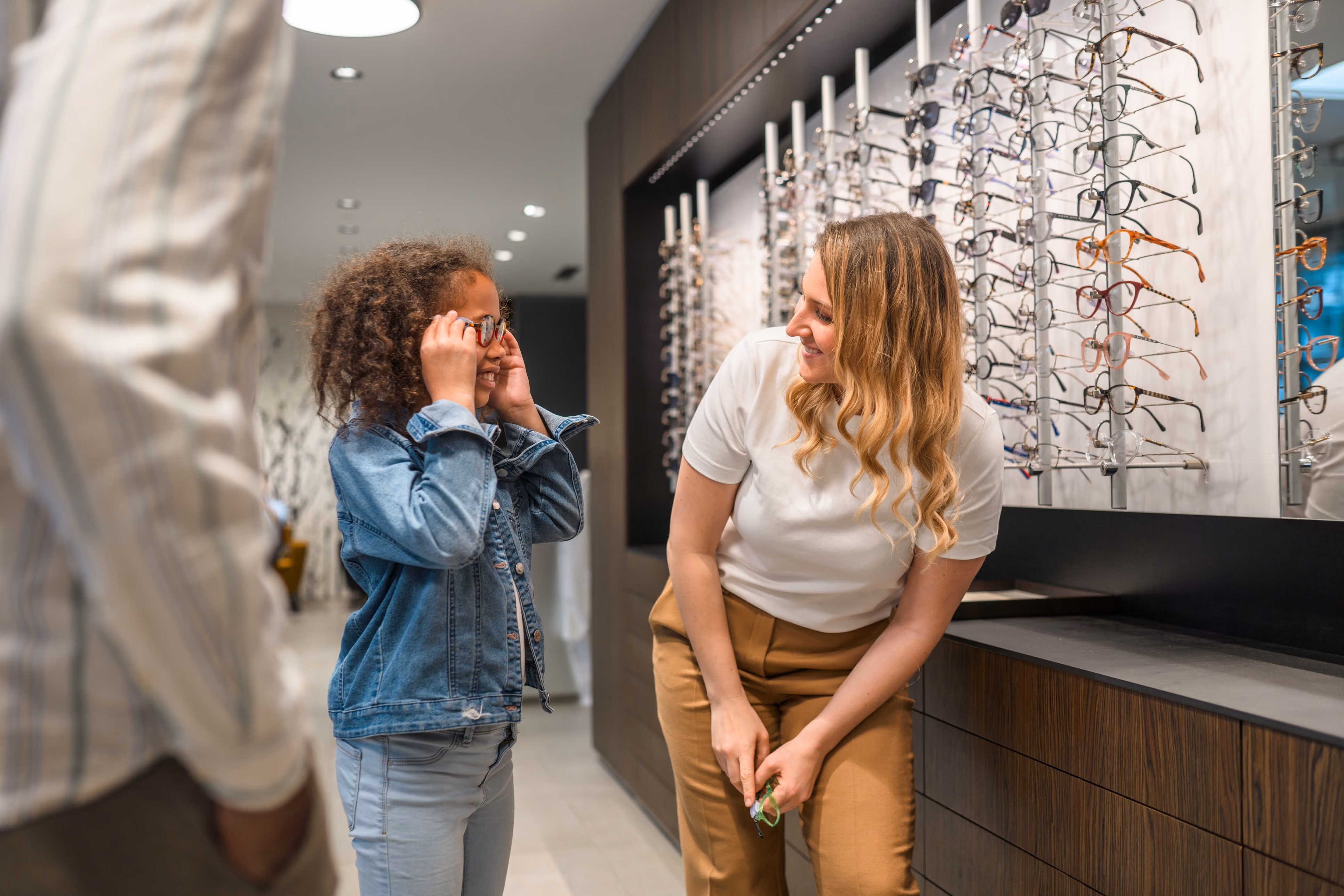 A young girl tries on glasses while a woman, likely a staff member, smiles and assists her near a display of various eyeglasses in an optical store.