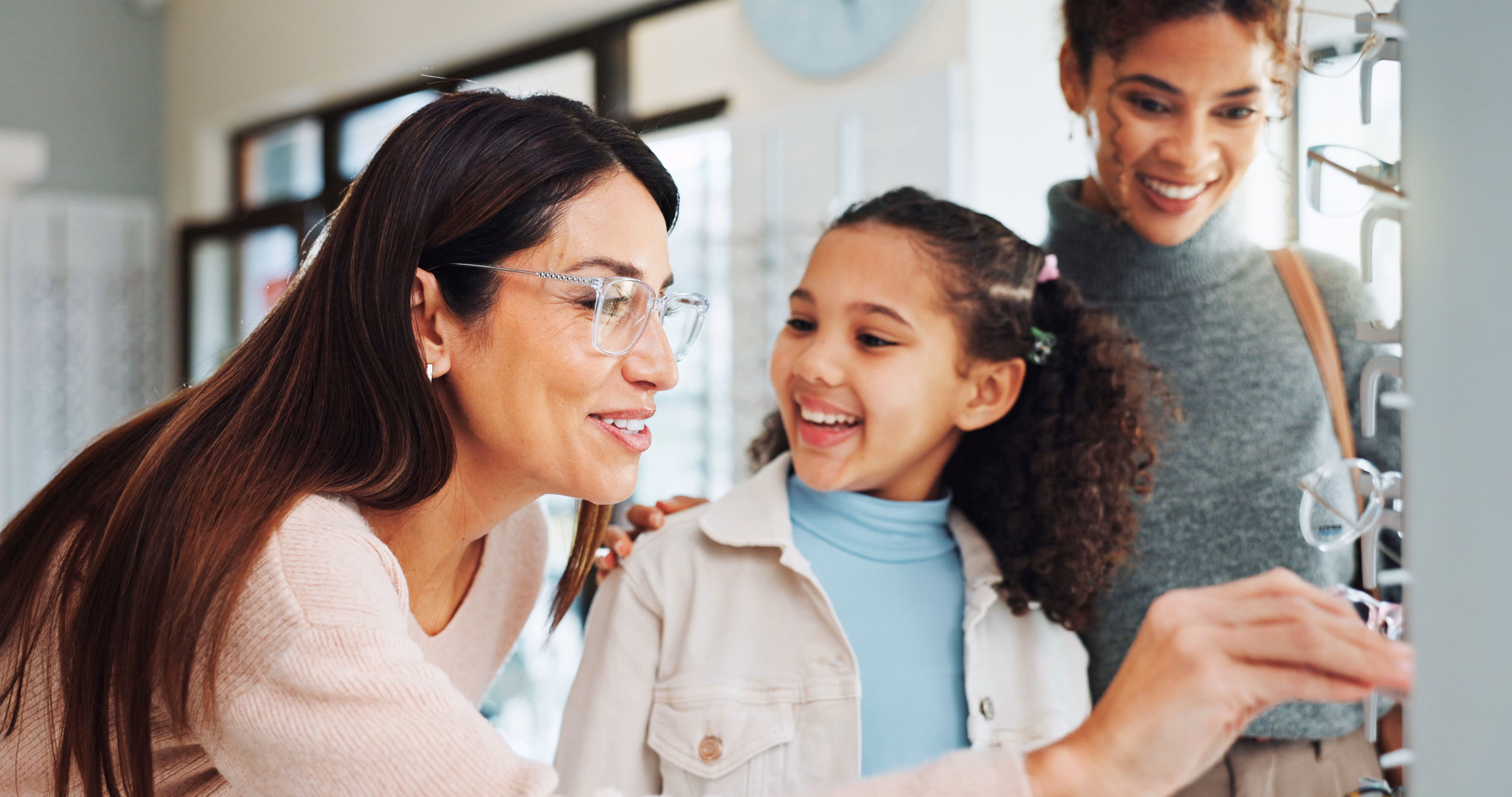 Two women and a young girl smiling while trying on eyeglasses at an optical store.