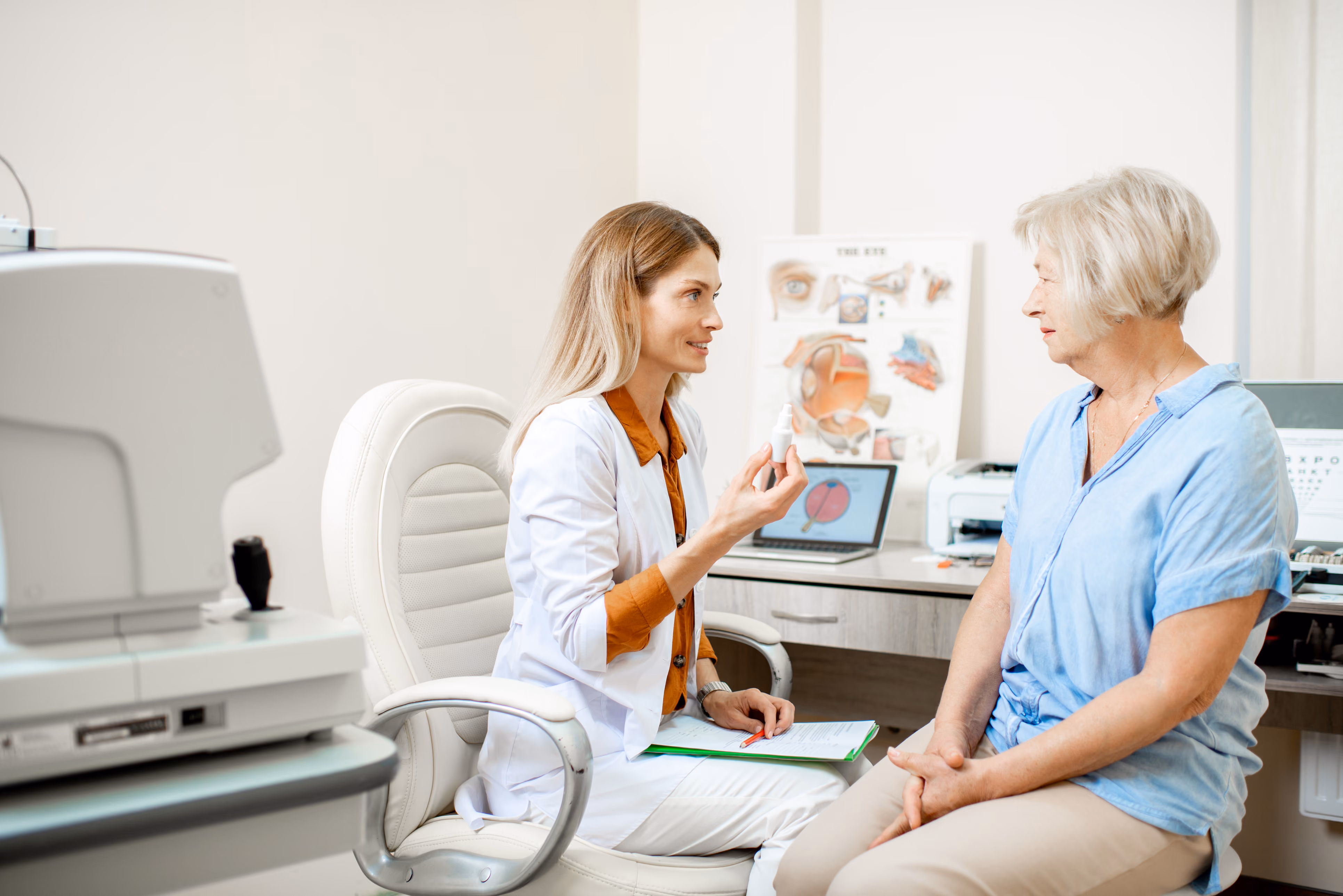 A female eye doctor in a white coat sitting on a chair and showing eye drops to an elderly female patient in a blue shirt inside an eye clinic.