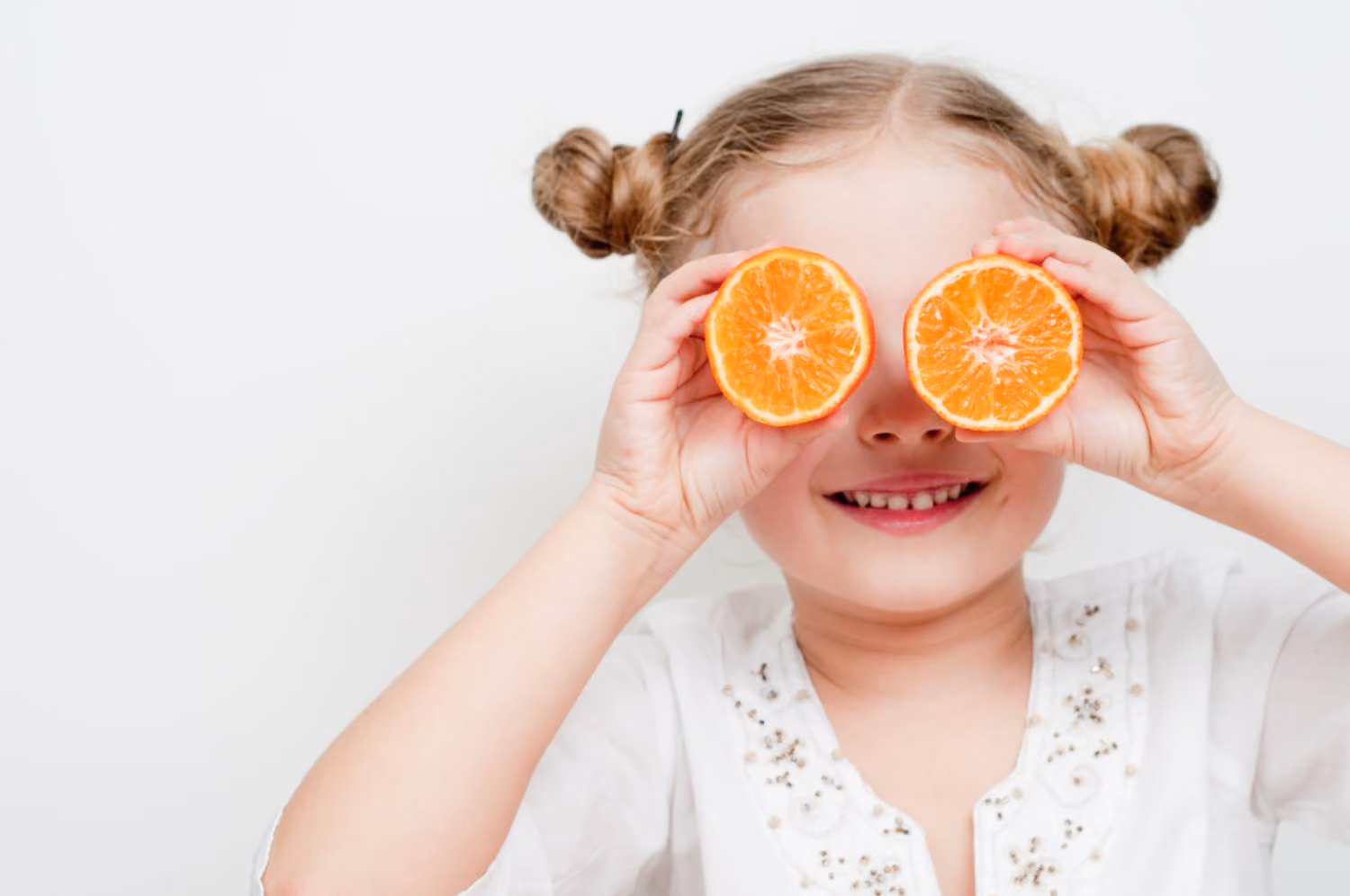 Young girl smiling while holding two orange halves over her eyes, representing Vitamin C's role in supporting children's eye health.