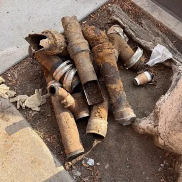 Rusty old broken drainage pipes with metal connectors on dirt near sidewalk and tree roots.