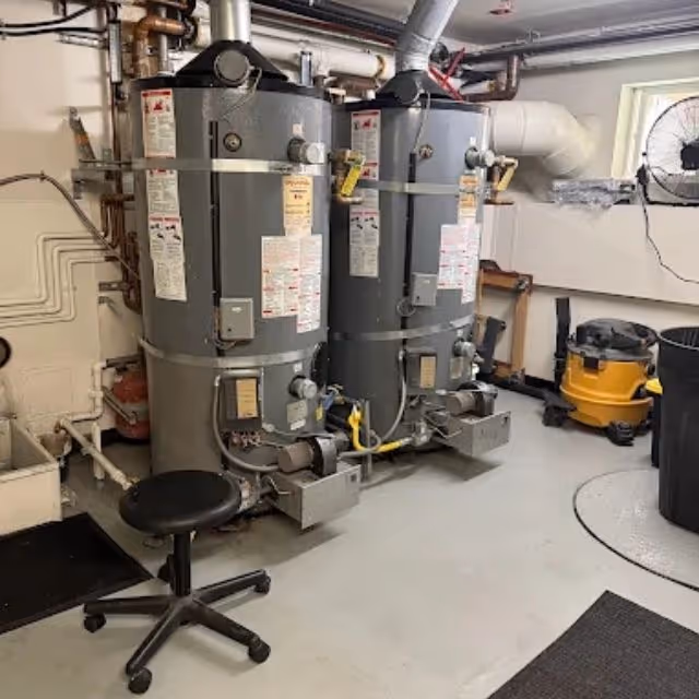 Two commercial water heaters installed in a utility room with gas and plumbing connections.