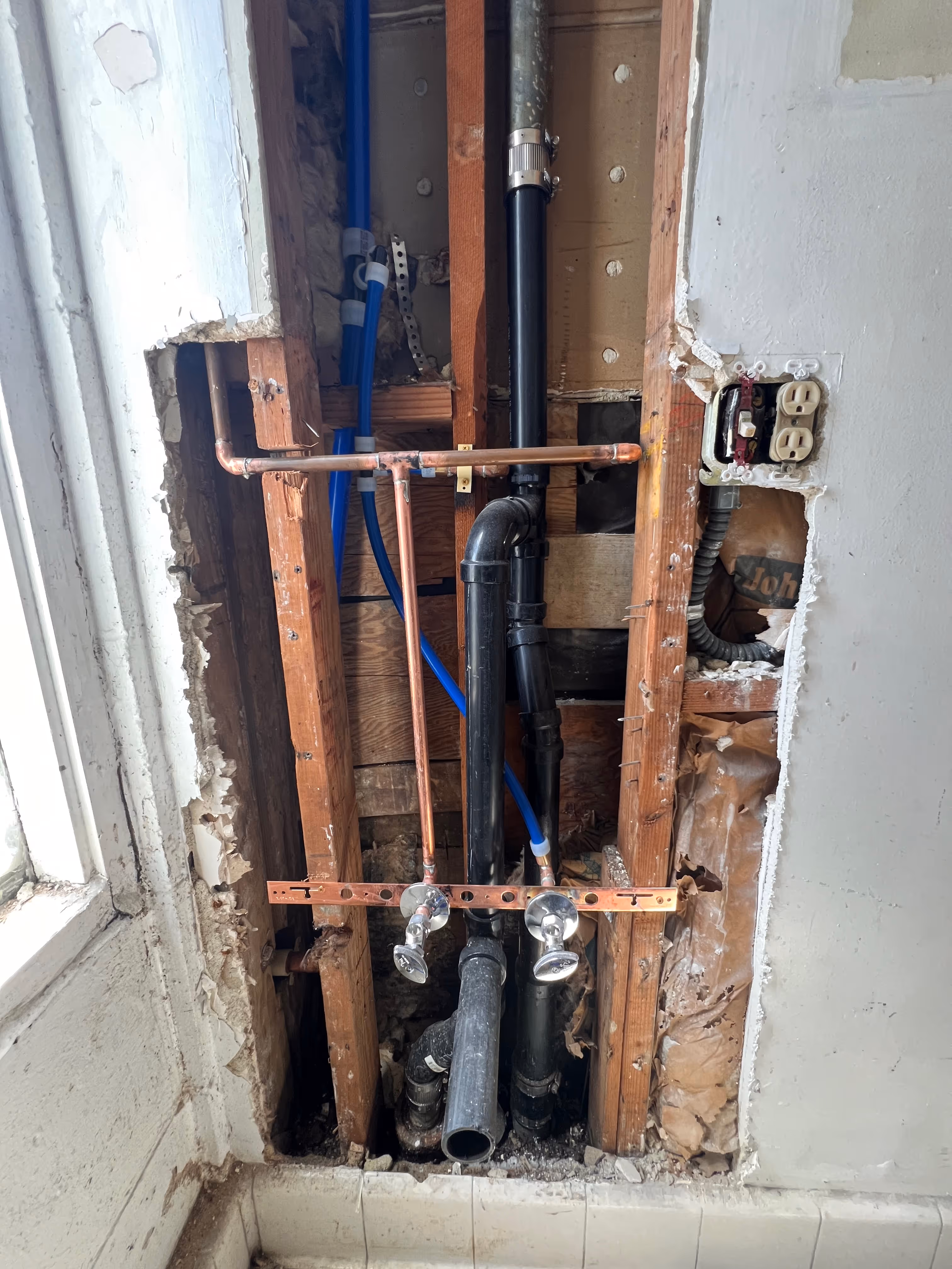Exposed wall plumbing with copper water lines and black drain pipe during bathroom rough-in installation.
