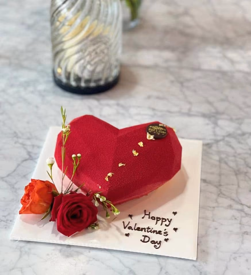 Red heart-shaped cake with gold flakes and flowers on white board saying Happy Valentine’s Day.