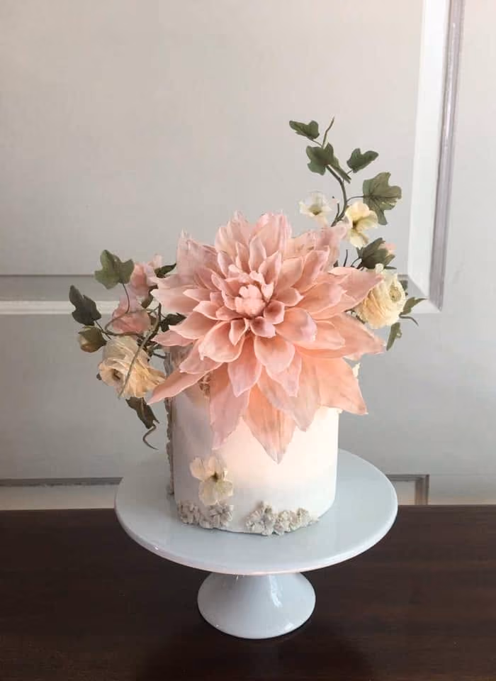 White cake with large pale pink flower and smaller white flowers on white cake stand.