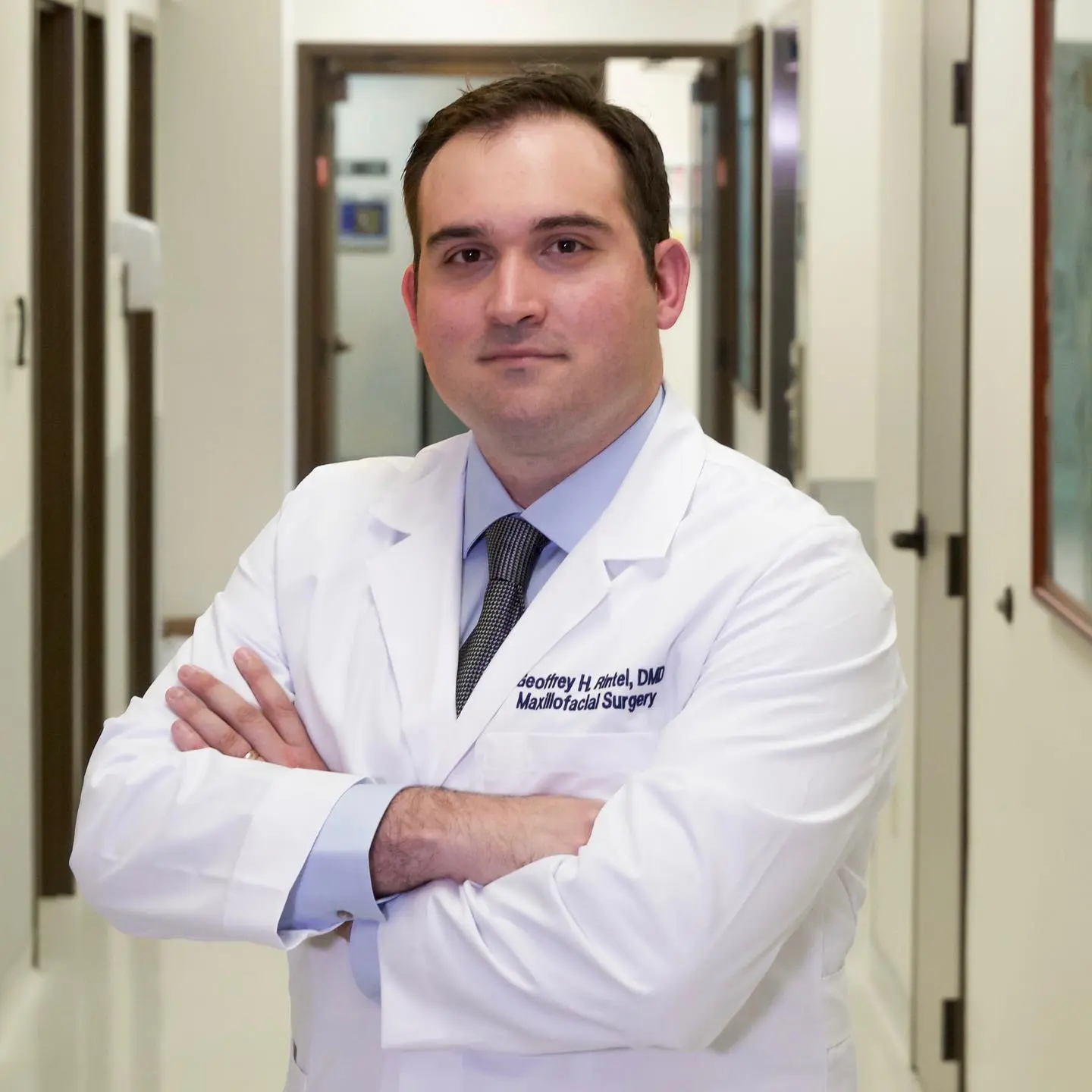 Man in a white lab coat and tie standing with arms crossed in a medical hallway.