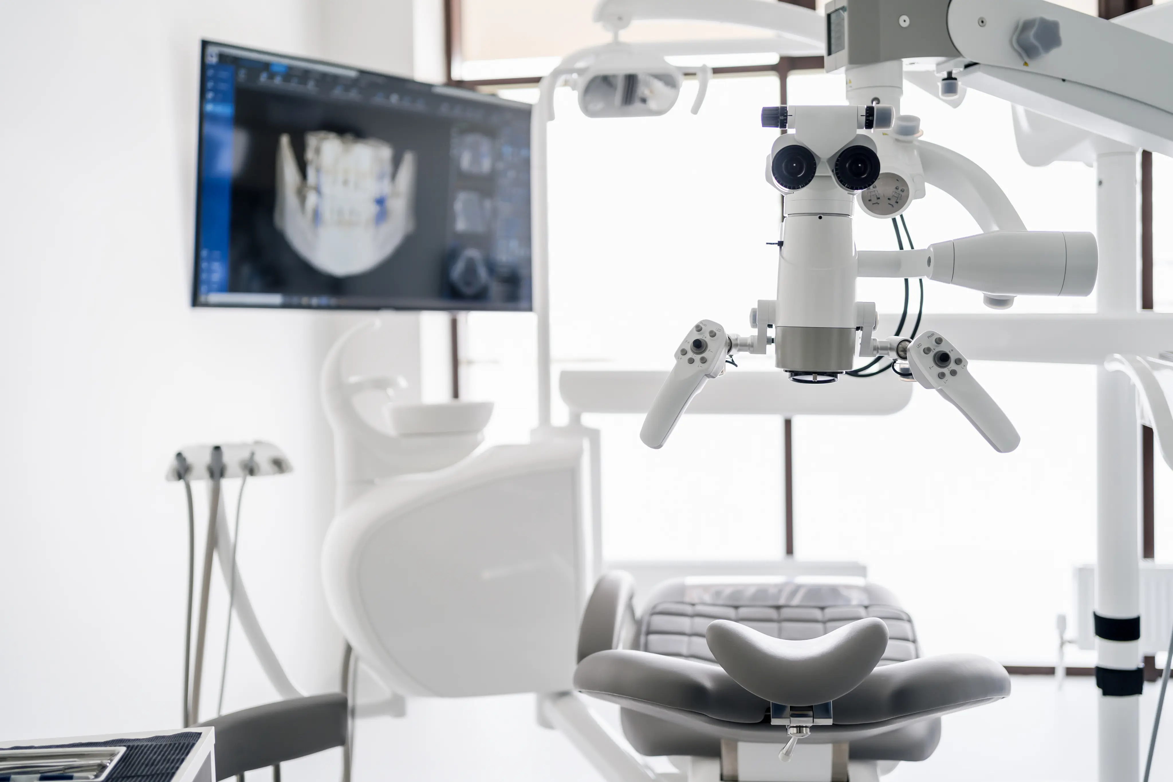 Modern dental examination chair with overhead dental microscope and a screen displaying a digital dental X-ray.