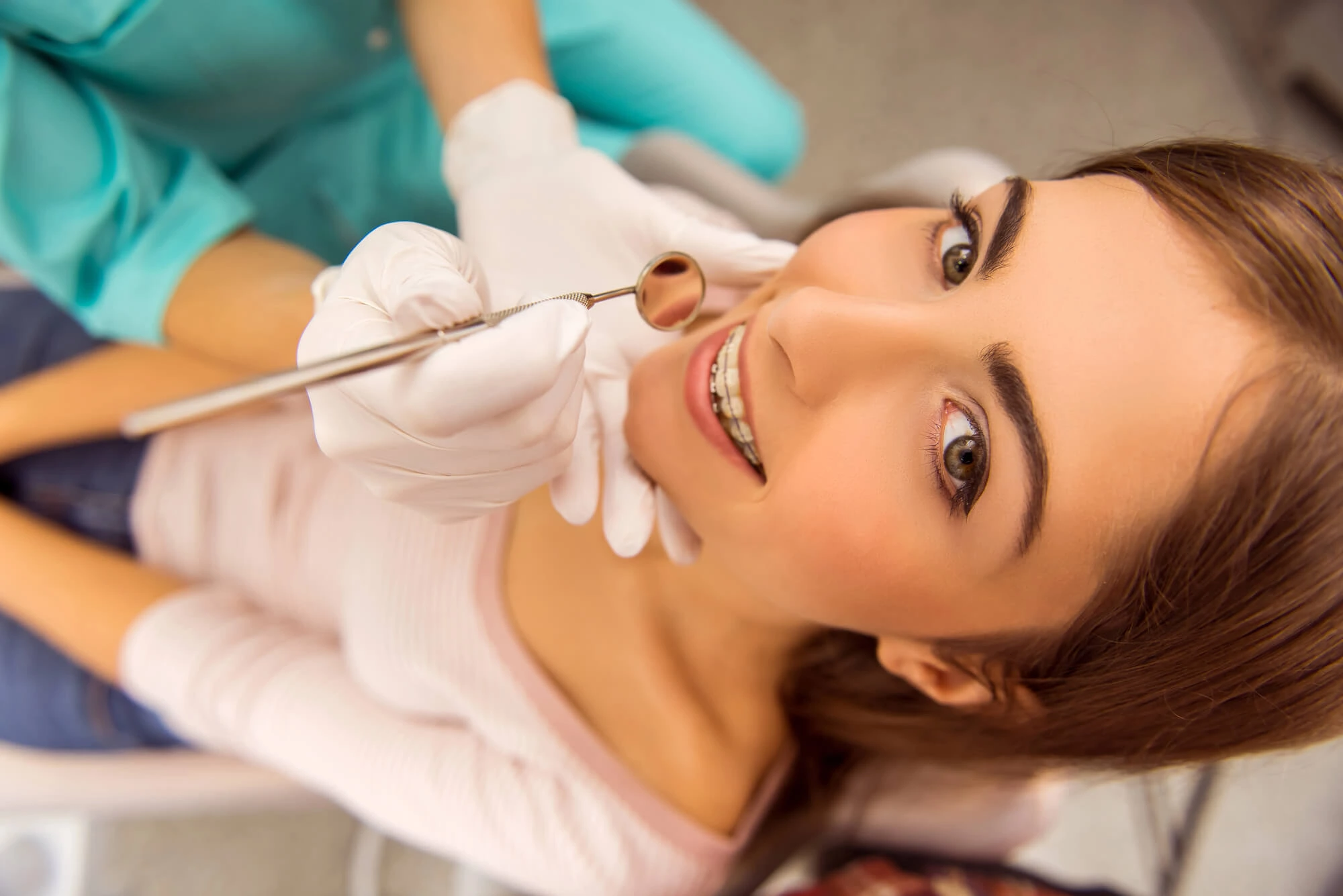 Smiling young woman with braces in a dental chair during a checkup with a dentist holding a dental mirror.