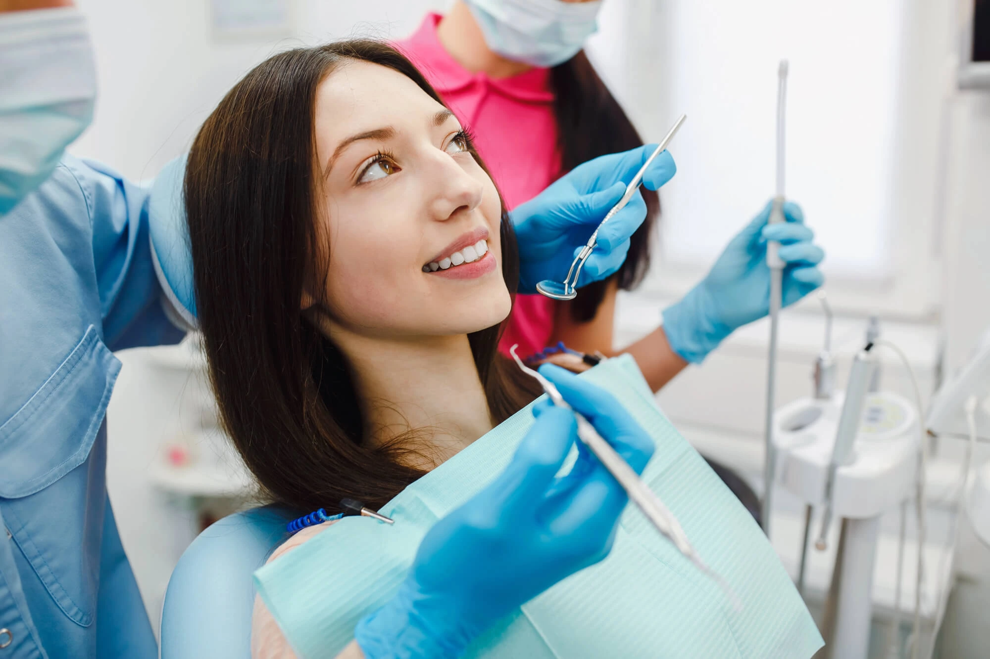 Young woman smiling in a dental chair while a dentist wearing blue gloves holds dental tools near her mouth.