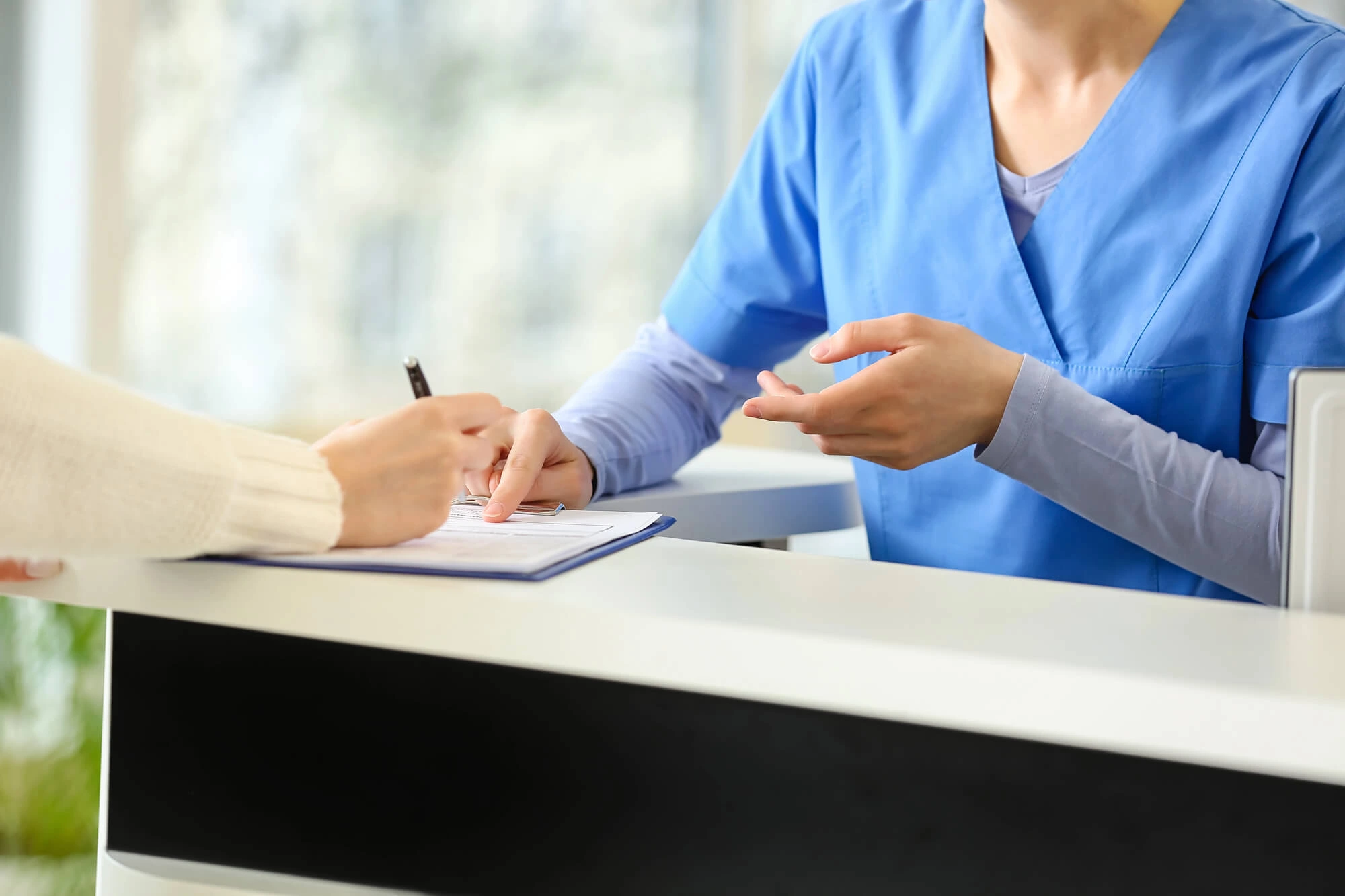 A healthcare professional in blue scrubs assisting a patient who is filling out a form on a clipboard at a reception desk.